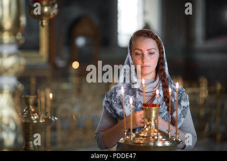Russian beautiful Caucasian woman with red hair and a scarf on her head is in the Orthodox Church, lights a candle and prays in front of the icon Stock Photo