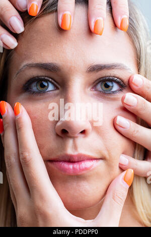 Attractive blond woman wearing makeup with female hands caressing her face in a close up conceptual image as she looks at the camera with a serious ex Stock Photo