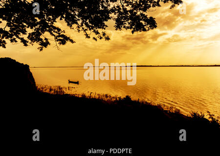 A lone fisherman moves out on a lake in a small boat as the sun breaks through evening Stock Photo