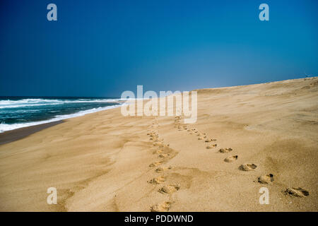 Two sets of footprints in sand , on a sandy beach disappearing into the distance with the breaking waves under a blue sky Stock Photo