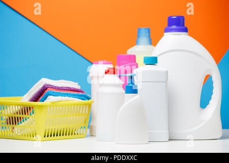 Photo of several bottles of cleaning products and multi-colored towels in basket isolated on blue, orange background Stock Photo
