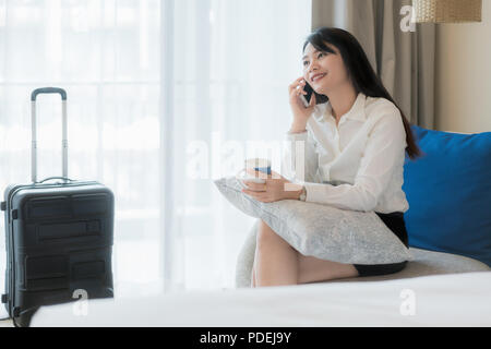 Beautiful Asian young smiling businesswoman in suit drinking coffee and using mobile phone while sitting on the sofa in hotel room. Business travel. Stock Photo