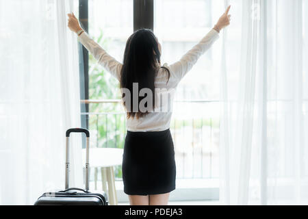 Young Asian business woman opens the window curtains and looks at view when she arrive in room at hotel. Business travel. Stock Photo