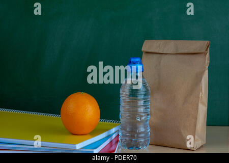 Lunch time. Brown paper lunch bag, orange and bottle of water on the desk with notebooks Stock Photo