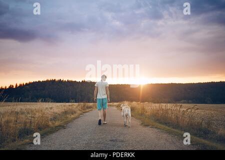 Rear view of the young man walking with his dog (labrador retriever) on the rural road at sunset.