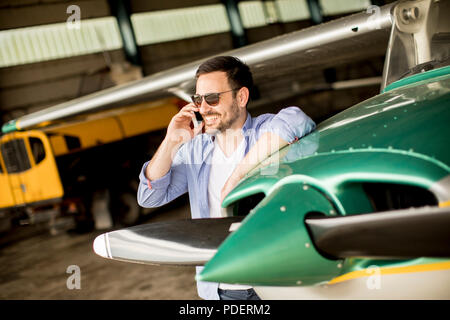 Young handsome young pilot checking airplane in the hangar and using mobile phone Stock Photo