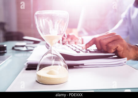 Close-up Of Hourglass In Front Of Businessperson's Hand Calculating Invoice Using Calculator Stock Photo