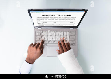 Overhead View Of A Businessman With Bandage Hand Filling Insurance Claim Form On Laptop Stock Photo