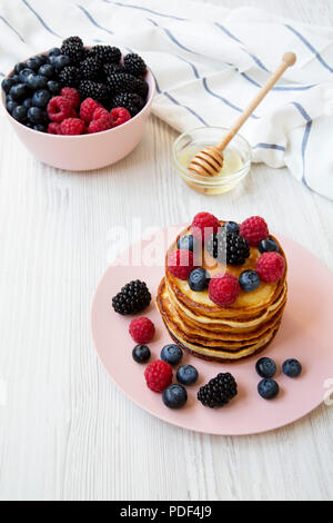 Pancakes with berries and honey on a pink plate over white wooden surface, high angle. Stock Photo