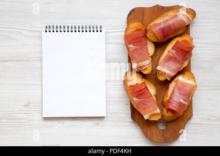 Crostini with serrano ham on rustic wooden board over white wooden background, top view. Blank notepad. Copy space. Stock Photo