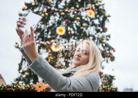 A beautiful young blonde woman or girl doing selfie or photographing next to a Christmas tree during Christmas holidays at the Old Town Square in Prague, Czech Republic. Stock Photo