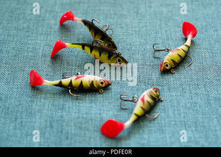 Silicone bait. Twisters on the background of burlap. Lures with triple hooks in the form of a small perch. Red tail. Stock Photo