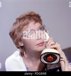 1950s glasses and telephone. A young woman in typical fifties glasses and bows is talking on the telephone.  Swedish on-piece plastic telephone created by Ericsson Company of Sweden. Because of its styling and its influence on future telephone design, the Ericofon is considered one of the most significant industrial designs of the 20th century. The idea was to incorporate the dial and handset into one single unit. The model is also known as the Cobra telephone for its resemblance to a coiled snake, and is now a sought after antique. Stock Photo