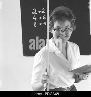 1950s schoolteacher. A young woman poses as a math teacher and is standing in front of the blackboard where a few figures are written. She smiles and is holding a wooden pointer in one hand and a school book in the other. 1958 Stock Photo