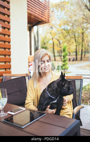Beautiful and happy blonde woman with tablet enjoying in cafe bar with her adorable French bulldog. Stock Photo