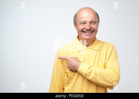 Happy senior man in yellow tshirt looking at camera, smiling and pointing aside with hand. Copy space for text. Stock Photo