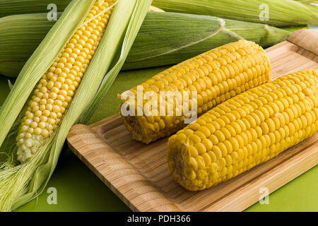 Fresh corn on the cob with leaves and boiled corn in wooden tray on green background Stock Photo