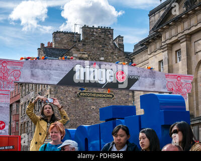 Edinburgh, Scotland, UK. 9th August 2018. Edinburgh Fringe Festival, Royal Mile, Edinburgh, Scotland, United Kingdom. On a sunny festival day the Virgin Money sponsored street festival is packed with people and fringe performers. An Asian woman tourist takes a selfie at the entrance to the Fringe street venue Stock Photo