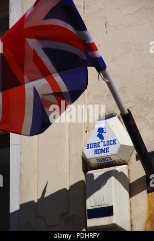 Sure Lock Home, Sherlock Holmes Burglar Alarm on a Building by a Union Jack Flag. Sidmouth, East Devon, UK. August, 2018. Stock Photo