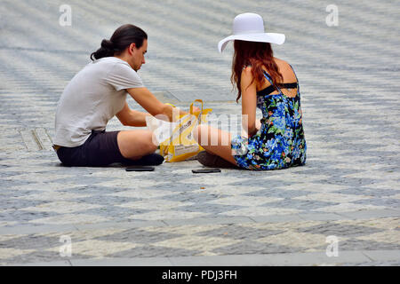 Young couple sitting on cobblestone pavement in square snacking on lunch, enjoying sunshine Stock Photo