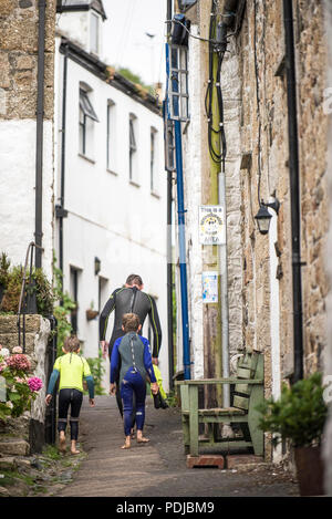 A man and two childrn wearing wetsuits walking up Duck street in Mousehole in Cornwall. Stock Photo