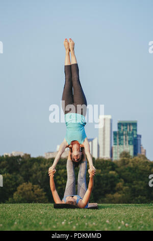 Two young Caucasian women yogi doing star shoulder stand acro yoga pose. Women doing stretching workout in park outdoors at sunset. Healthy lifestyle  Stock Photo