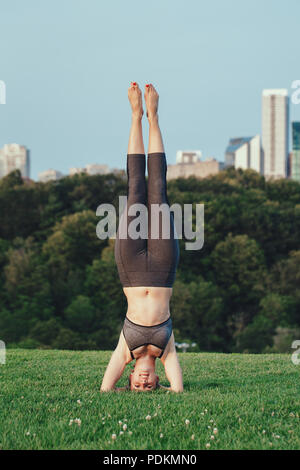 Young beautiful Caucasian woman doing headstand yoga pose. Yogi woman doing stretching workout in park outdoors at sunset. Healthy lifestyle modern ac Stock Photo