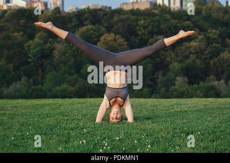 Young beautiful Caucasian woman doing headstand yoga pose. Yogi woman doing stretching workout in park outdoors at sunset. Healthy lifestyle modern ac Stock Photo