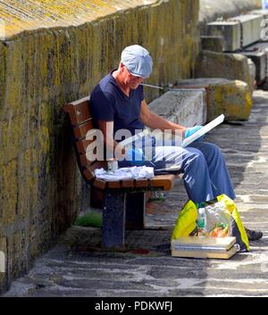 Senior retired artist sitting on a bench painting,Mousehole,Cornwall,England,UK Stock Photo