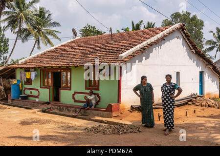 Nilavagilukaval, Karnataka, India - November 1, 2013: Two smiling woman and a man in front of their one story stone building with red pan roof. Brown  Stock Photo