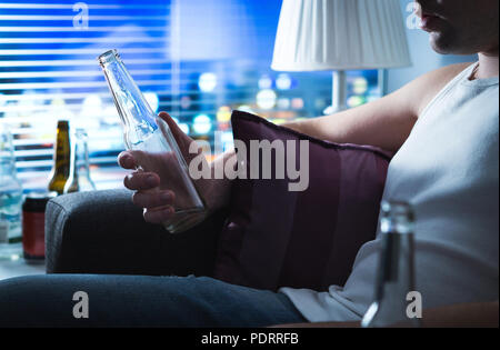 Drunk man sitting on sofa and holding a bottle of alcohol. Alcoholic drinking home on couch late at night. Alcoholism, alcohol abuse and depression. Stock Photo