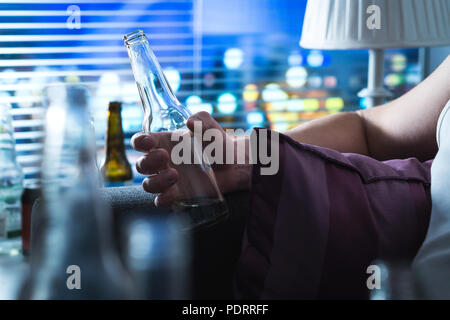 Man with drinking problem late at night. Alcoholic sitting on sofa. Drunken person holding a bottle. Alcoholism and alcohol abuse concept. Stock Photo