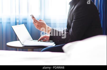 Business man in hotel room with smartphone and laptop. Businessman reading emails or using social media. Remote work during business trip. Stock Photo