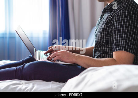 Man using laptop in hotel room or home bedroom. Guy lying on bed and typing with notebook computer. Person in the morning with modern device. Stock Photo