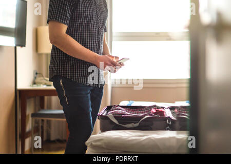 Man using smartphone in hotel room with open baggage and suitcase on bed. Mobile phone and packing. Just arrived or leaving. Stock Photo