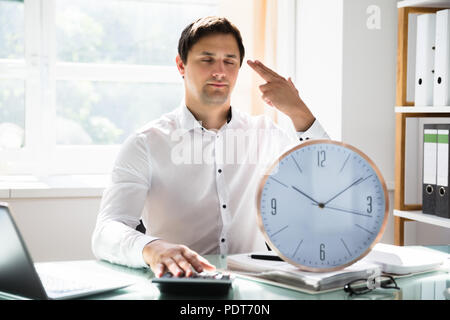 Tired young businessman making shooting gesture behind clock in office Stock Photo