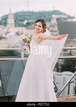 Smiling bride is correcting the veil while standing near the balcony fence. Stock Photo