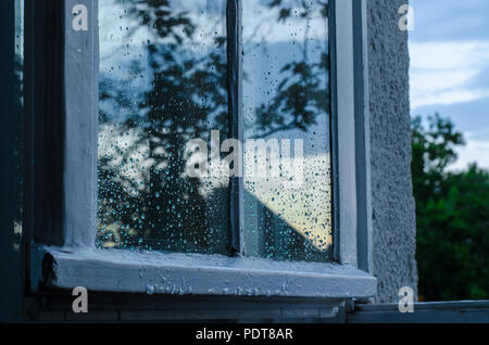 Window covered with raindrops after a summer rain, showing a reflection of the outside nature and sky basked in blue light of the evening Stock Photo