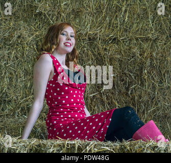 Red headed girl with red vintage polka dot dress and pink wellington boots sits on hay bales in a cosy barn. Stock Photo