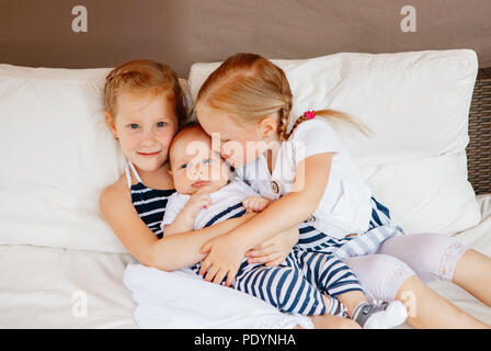 Lifestyle portrait of cute white Caucasian girls sisters holding kissing little baby, sitting on bed indoors. Older siblings with younger brother sist Stock Photo