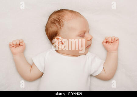 Portrait of sleeping cute Caucasian little baby newborn in white clothes lying on bed with his hands up. Lifestyle candid real concept. Stock Photo