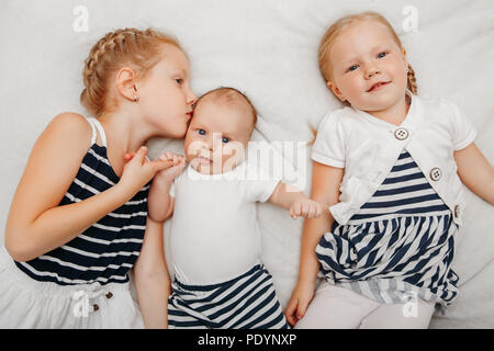 Lifestyle portrait of cute white Caucasian girls sisters holding kissing little baby, lying on bed indoors. Older siblings with younger brother sister Stock Photo