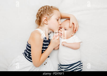 Lifestyle portrait of cute white Caucasian girl sister holding kissing little baby, lying on bed indoors. Older sibling with younger brother newborn.  Stock Photo