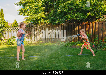Two girls splashing each other with gardening house on backyard on summer day. Children playing with water outside at sunset. Candid moment, lifestyle Stock Photo