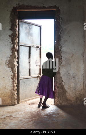 Young Ugandan girl stands in the doorway of her school classrom looking out at other pupils, wearing a bright coloured skirt Stock Photo