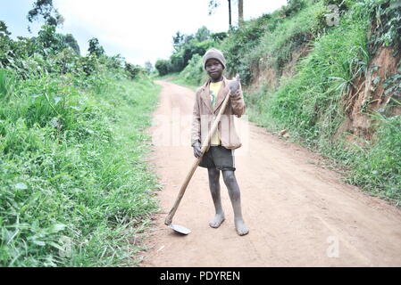 Young Ugandan boy stands in the middle of a dirt road holding farm tools, barefoot, looking at the camera Stock Photo