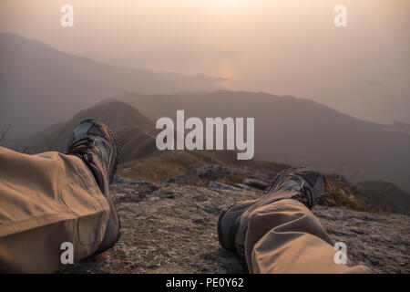 feet and leg of hiker sit on peak with hiking trail and mountain range background with fog at sunrise in winter, Lantau Peak, Hong Kong Stock Photo