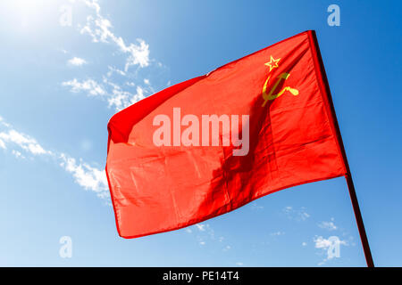 Red communist flag of the USSR against the blue sky with white clouds and sun. Stock Photo