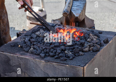 Red hot metal being forged in a blacksmiths fire in a smith next to an anvil. The blacksmith is holding tongs in the fire which is glowing red hot Stock Photo