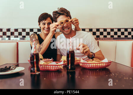 Woman sitting at a restaurant with man and having fun holding biscuit rings to his eyes. Romantic couple at a diner sitting with food and soft drinks Stock Photo